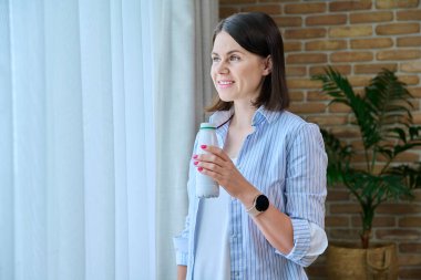 Evde, pencerenin yanında, şişeden mayalanmış süt içen genç bir kadın sağlıklı bir kahvaltı yapıyor. Süt ürünleri, yoğurt, kefir, diyet, sağlıklı beslenme konsepti