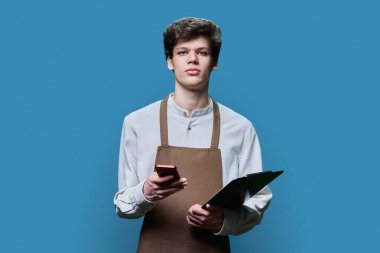 Önlüklü, elinde kağıt pano, elinde akıllı telefon olan mavi stüdyo arka planındaki kameraya bakan genç bir adamın portresi. Hizmet, sipariş, çevrimiçi düzen, küçük işletme, personel, meslek