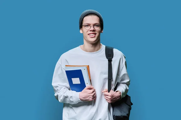年轻的大学生背着背包 拿着笔记本课本 头戴针织帽眼镜 身穿白色运动衫 在蓝色的工作室背景下看着相机 学习概念 — 图库照片