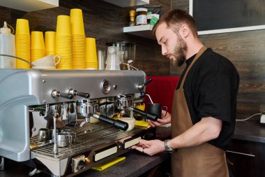 Kahve dükkanında kahve hazırlayan erkek barmen, kahve makinesinin yanında duran önlüklü, sakallı genç adam. Küçük işletme, hizmet, kahve dükkanı, personel, iş kavramı