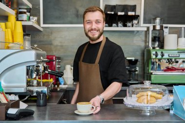 Önlüklü genç barista erkek portresi. Yeni hazırlanmış cappuccino kahvesi. Kahve dükkanında tezgahın arkasında duruyor. Küçük işletme hizmeti, kafe, personel iş konsepti
