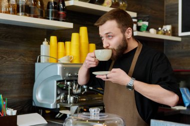 Önlüklü genç barista erkek portresi. Yeni hazırlanmış cappuccino kahvesi. Kahve dükkanında tezgahın arkasında duruyor. Küçük işletme hizmeti, kafe, personel iş konsepti