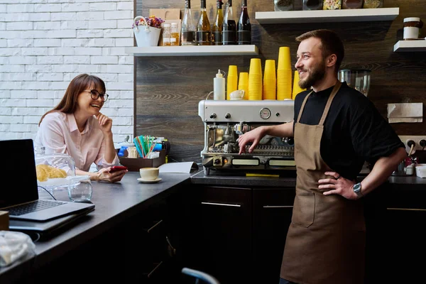 年轻的男性咖啡师在咖啡店里与女顾客交谈 旁边的柜台上放着一杯刚准备好的卡布奇诺咖啡 小企业 服务业 咖啡店 工作概念 — 图库照片