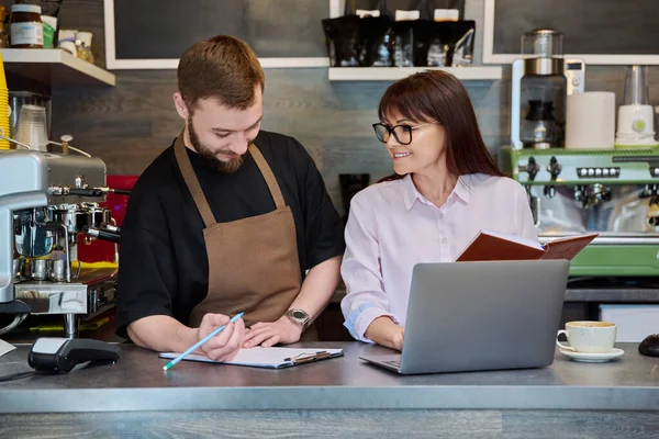 同僚のパートナー 若い男性と成熟した女性がコーヒーショップのバーの後ろに立ってラップトップを使用して話をしています チーム 小企業 スタッフ カフェカフェテリアレストラン 起業家精神 — ストック写真