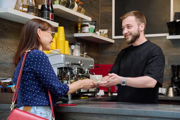 Young male barista talking to woman customer in coffee shop, near counter with cup of freshly prepared cappuccino coffee. Small business, service, coffee shop, staff, work concept