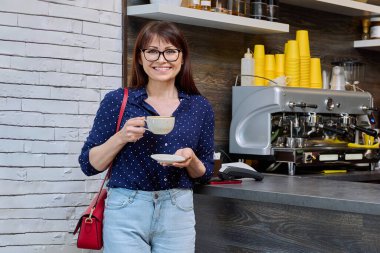 Tezgahın yanındaki kameraya bakan bir fincan taze kokulu kahveyle olgun bir bayan kahve müşterisi. Kahve içecekler, yemek, satış, servis, dinlenme konsepti