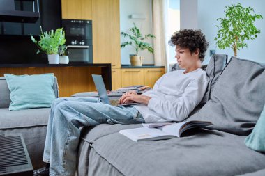 Yakışıklı bir üniversite öğrencisi, evde çalışıyor, kanepede oturuyor ve dizüstü bilgisayarını daktilo ediyor. Genç erkek, 18, 19 yaşında, oturma odasının içinde kıvırcık saçlı. Eğitim bilgileri çevrimiçi internet teknolojisi