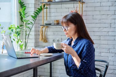 Orta yaşlı bir kadın, kahve içiyor, laptop ekranına bakıyor, kafede masada oturuyor, kahve dükkanında. Uzaktan çalışma, serbest çalışma, blog yazma, sohbet, online internet teknolojisi, olgun insanlar konsepti