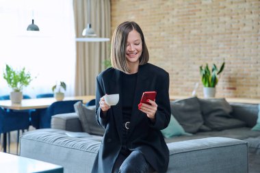 Oturma odasındaki kanepede elinde bir fincan kahveyle akıllı telefon kullanan şık, genç ve güzel bir kadın. İnternet teknolojisi, gezici uygulamalar, boş zaman işi alışverişi.