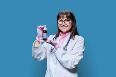 Kadın doktor, mavi arka planda koyu kahverengi ilaç şişesi gösteriyor. Beyaz önlüklü yetişkin bir kadın kameraya bakıyor. Eczacılık İlaç İlaç Tedavisi