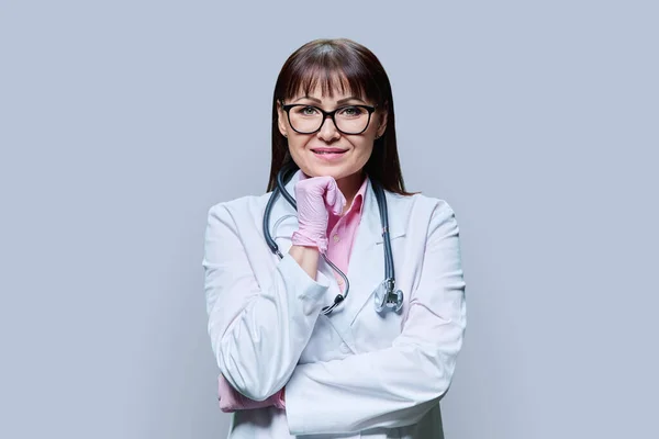 白色实验室外套手套听诊器中中年女医生的画像 在灰色工作室背景下看着相机 医药健康保护 专业医务工作者 医疗服务概念 — 图库照片