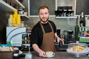 Önlüklü genç barista erkek portresi. Yeni hazırlanmış cappuccino kahvesi. Kahve dükkanında tezgahın arkasında duruyor. Küçük işletme hizmeti, kafe, personel iş konsepti