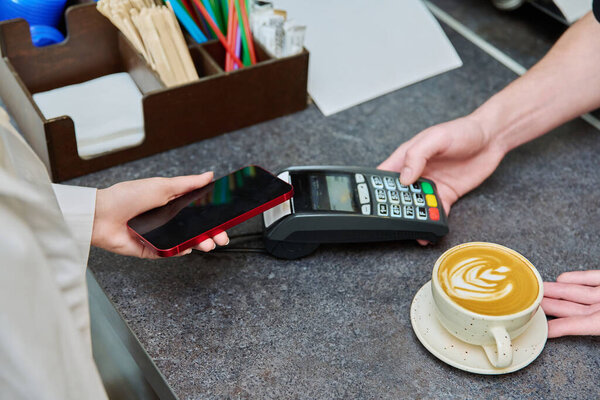 Close-up of a customers hand with a smartphone paying for a cup of coffee in a cafe, contactless using the terminal, mobile payment
