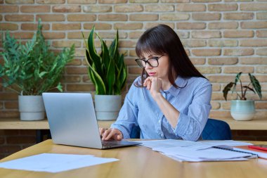 Orta yaşlı, kendine güvenen, ciddi bir iş kadını ofis çalışanı, işletme evraklarıyla çalışan, masa başında oturan bir yönetici. İş, iş, insanlar, İnternet teknolojisi