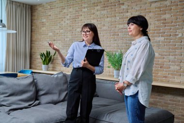 Kendine güvenen kadın emlakçı, apartman dairesini kiracıya olgun bir kadın olarak gösteriyor. Mobilyalı modern oturma odasını gösteren panosu olan kadın emlakçı. Satılık ev kiralama işi