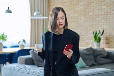 Oturma odasındaki kanepede elinde bir fincan kahveyle akıllı telefon kullanan şık, genç ve güzel bir kadın. İnternet teknolojisi, gezici uygulamalar, boş zaman işi alışverişi.