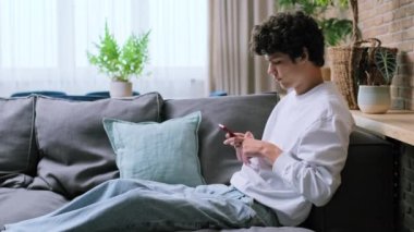 Oturma odasındaki kanepede oturan akıllı telefonu kullanan genç yakışıklı adam. Erkek, 20 yaşında, kıvırcık saçlı. Boş zaman iletişimi için internet çevrimiçi mobil uygulamaları