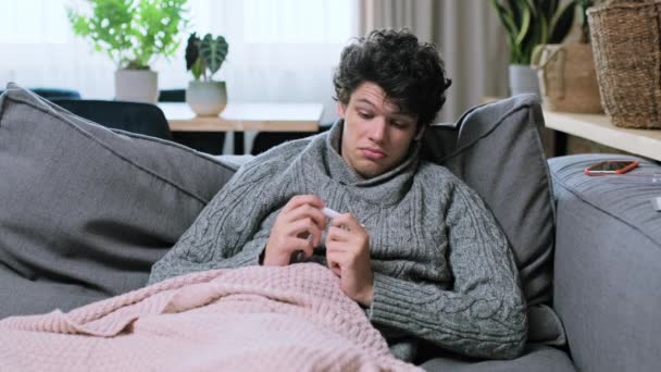Smutny Chory Młodzieniec Patrzący Temperaturę Termometrze Ubrany Wełniany Sweter Leżący — Wideo stockowe