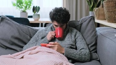 Kareli gömleğinin altında yün süveter giyen genç bir erkek evinde sıcak bir bardak sıcak içecekle kendini ısıtıyor, akıllı telefon kullanarak rahatlıyor. Sonbahar, kış, ısıtma mevsimi, soğuk mevsim