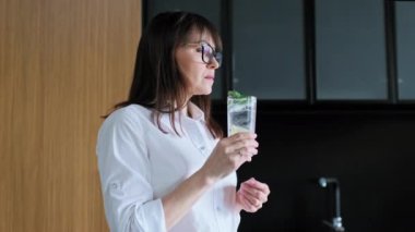 Olgun bir kadın limonlu nane şekerli bir bardak su içiyor, evde mutfakta duruyor. Sıcaklık yaz gününde diniyor, sağlıklı diyet içeceği.