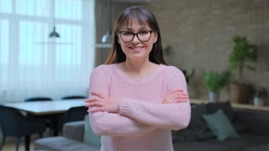Evde kendinden emin orta yaşlı bir kadının portresi. Kollarını kavuşturup gülümseyen başarılı kadın kameraya bakıyor, oturma odası arka planı.
