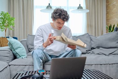 Genç bir erkek online alışveriş paketini açıyor, evdeki koltukta yeni akıllı telefonlu bir kutuyla oturuyor. İnternet alışverişi, dağıtım, gençlik kavramı