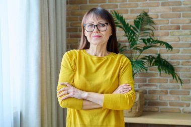 Evde kendinden emin orta yaşlı bir kadının portresi. Sarı elbiseli, kollarını kavuşturup kameraya bakan başarılı bir kadın.