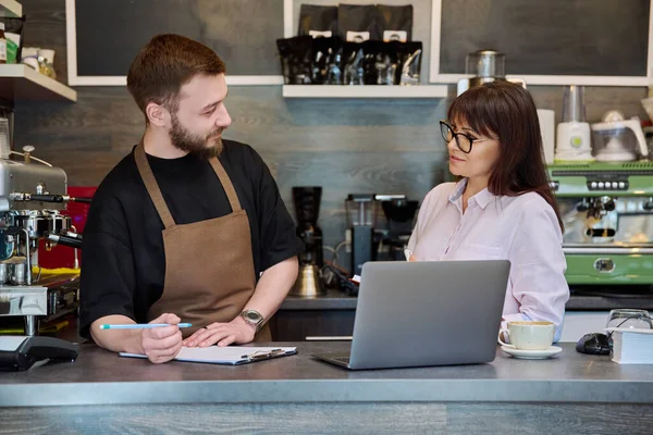 同僚のパートナー 若い男性と成熟した女性がコーヒーショップのバーの後ろに立ってラップトップを使用して話をしています チーム 小規模ビジネス スタッフ チームワーク 起業家精神 パートナーシップの概念 — ストック写真