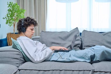 18-20 yaşlarındaki genç adam evde dinleniyor, oturma odasında kanepede uzanıyor, kulaklıkla dizüstü bilgisayara bakıyor. İnternet teknolojisi, eğlence iletişim kavramı