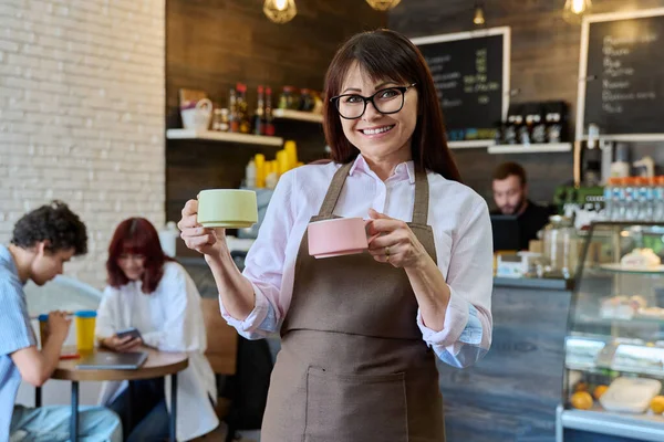 2杯のコーヒーを手にした笑顔の女性コーヒーショップ労働者の姿 カフェホール内のカメラを見てエプロンの女性 小規模ビジネス フードサービスの職業 スタッフ ワークコンセプト — ストック写真