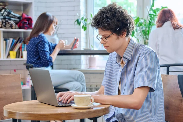 英俊的卷曲小伙子大学生坐在咖啡店的桌旁 一边喝着咖啡 一边在笔记本电脑上打字 用于休闲交流学习 青年生活方式的互联网在线技术 — 图库照片