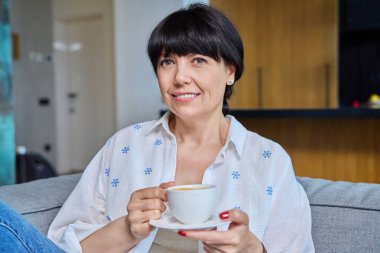 Gülümseyen olgun bir kadın evde bir fincan kahve ile rahatlıyor. Oturma odasında koltukta oturan, kameraya bakan 50 'li yaşlarda pozitif bir kadın. Yaşlılar, yaşam tarzı, güzellik, sağlık konsepti