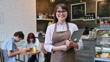 Kendine güvenen orta yaşlı kadın kahve dükkanı sahibinin portresi. Önlüklü bir kadın dizüstü bilgisayarını tutuyor, kafenin içindeki kameraya bakıyor. Küçük işletme, gıda hizmeti işi, insanlar, personel, iş, iş kavramı