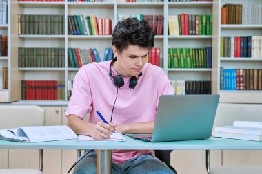 Üniversite kütüphanesinde dizüstü bilgisayar kitaplarıyla oturan genç bir erkek öğrenci. 18-20 yaşlarında, kulaklıklı, defterine yazı yazan bir adam. Bilgi, eğitim, gençlik, üniversite konsepti