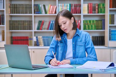Kütüphanede okuyan genç bir kız öğrenci, bilgisayar laptopu kullanıyor, deftere yazıyor. Bilgi, eğitim, gençlik, üniversite konsepti