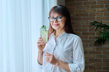 Olgun bir kadın, antioksidanlı içecekle, limonlu buzlu köpüklü su, pencerenin yanındaki evde. Sağlıklı yaşam tarzı, yiyecek ve içecek, diyet, beslenme konsepti