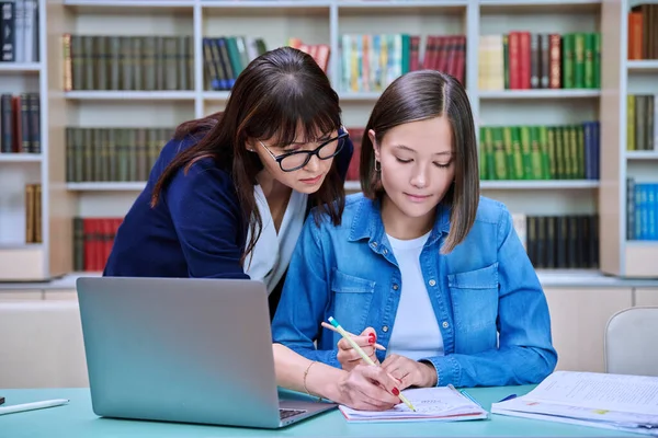 Young female university student with middle aged teacher mentor preparing for exam sitting at desk with laptop books inside library. Knowledge, education, youth, college university concept