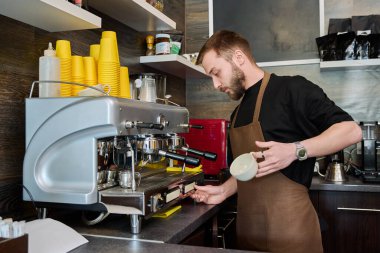 Kahve dükkanında kahve hazırlayan erkek barmen, kahve makinesinin yanında duran önlüklü, sakallı genç adam. Küçük işletme, yemek servisi işi, kahve dükkanı, personel, iş konsepti