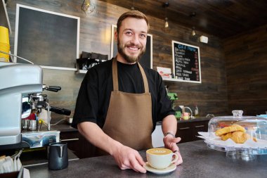 Önlüklü genç barista erkek portresi. Yeni hazırlanmış cappuccino kahvesi. Kahve dükkanında tezgahın arkasında duruyor. Küçük işletme hizmeti, kafe, personel iş konsepti