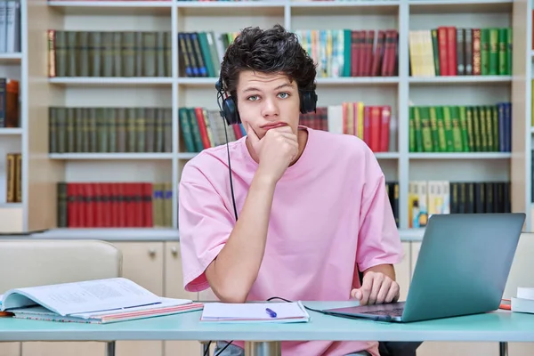 在大学图书馆里 年轻的男生坐在电脑笔记本电脑旁看书 20岁的家伙 带着耳机看着摄像机 大学概念 — 图库照片