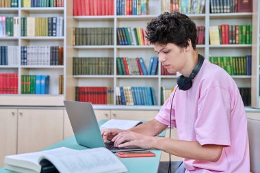 Üniversite kütüphanesinde dizüstü bilgisayarla oturan genç bir erkek öğrenci. 18-20 yaşlarında, kulaklıklı, dizüstü bilgisayarda yazan bir adam. Bilgi, eğitim, gençlik, üniversite konsepti