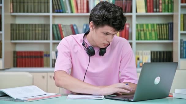 大学図書館にノートパソコンの本を持って座っている若い男子学生 20歳の男性は ヘッドフォンでノートブックを書いています 大学のコンセプト — ストック動画