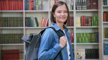 Sırt çantasıyla eğitim binasının kütüphanesinde kameraya bakan genç ve gülümseyen bir üniversite öğrencisi. Bilgi, eğitim, gençlik, üniversite konsepti