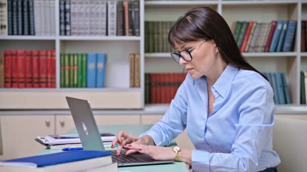 真剣な中年の女性は 図書館の中のラップトップで働いています 女性教師 メンター 教育者 レッスン準備講師 — ストック動画