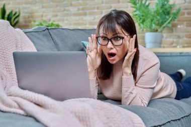 Şaşırmış orta yaşlı bir kadın oturma odasında kanepede dizüstü bilgisayar ekranına bakıyor. İnternet teknolojilerini kullanarak internet üzerinden alışveriş sitelerinde sohbet eden bir kadın.