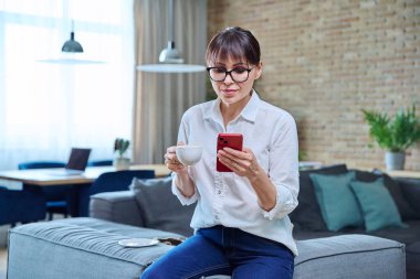 Orta yaşlı kadın akıllı telefon kullanıyor, evinde kanepede kahve içiyor. Olgun kadın dinlenme, okuma haberleri, sosyal ağlar, arkadaşlarla sohbet, iş arkadaşları, aile, mobil uygulamalar, boş zaman