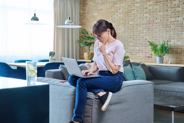 Serious middle aged woman using laptop while sitting on sofa at home. Thoughtful female looking at computer screen, solving difficulties and problems