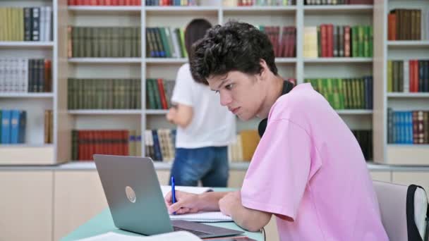 在大学图书馆里 年轻的男生坐在笔记本电脑前看书 18到20岁的家伙带着耳机在笔记本上写字知识 大学概念 — 图库视频影像