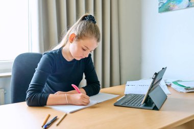 Kız çocuğu evde ders çalışıyor, masasında dijital tablet kullanıyor, deftere yazıyor, derse hazırlanıyor, sınava hazırlanıyor. Eğitim, bilgi, okul, çocukluk kavramı