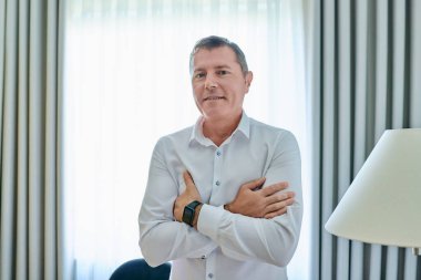 Kendine güvenen olgun bir adamın portresi. 50 yaşında, beyaz gömlekli, çapraz kollu kameraya bakıyor. Ofisin içinde poz veriyor.